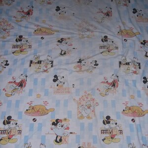 Pu&ograve; includere: Un lenzuolo in tessuto bianco con un motivo ripetuto di Topolino, Minnie, Paperino e Pluto in varie scene. La stampa include il testo Mickey's Country Store, con accenti rettangolari azzurri distribuiti su tutto il design.