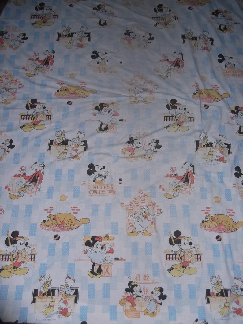 Pu&ograve; includere: Un lenzuolo in tessuto bianco con un motivo ripetuto di Topolino, Minnie, Paperino e Pluto in varie scene. La stampa include il testo Mickey's Country Store, con accenti rettangolari azzurri distribuiti su tutto il design.