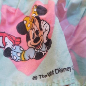 Puede incluir: Una tela rosa y verde con una imagen de dibujos animados de Minnie Mouse con un vestido azul con lunares blancos y un lazo rojo. La tela tiene el texto "The Walt Disney Company" en negro.