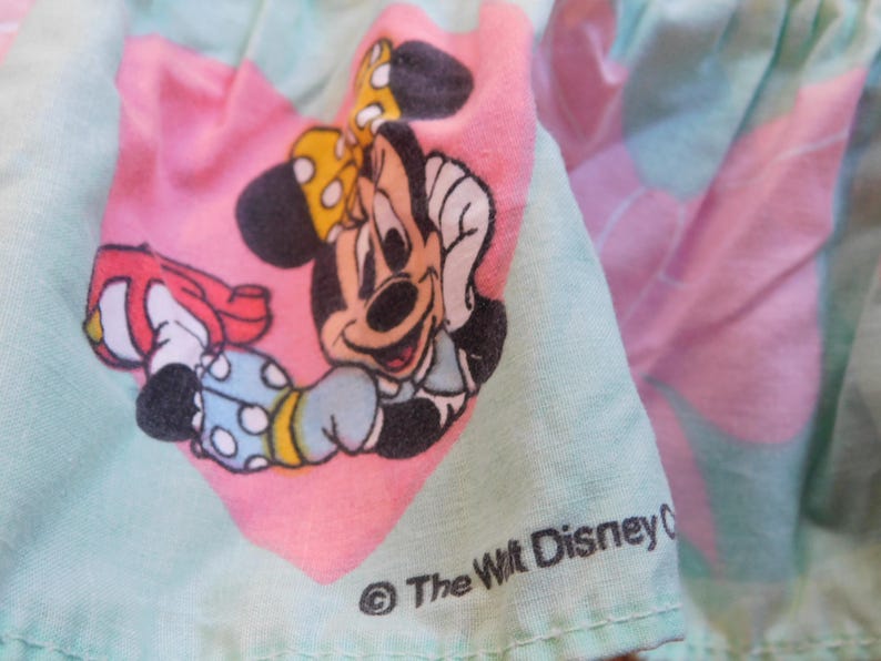 Puede incluir: Una tela rosa y verde con una imagen de dibujos animados de Minnie Mouse con un vestido azul con lunares blancos y un lazo rojo. La tela tiene el texto "The Walt Disney Company" en negro.