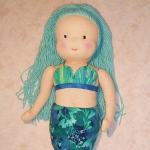May include: A handmade doll with long turquoise yarn hair, wearing a green and blue patterned swimsuit top and a green and blue floral print mermaid tail.