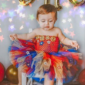 May include: A young child wearing a red, blue, and gold tulle tutu with stars and a red, white, and blue patterned bodice. The child is sitting on a white stool with a blue cushion.