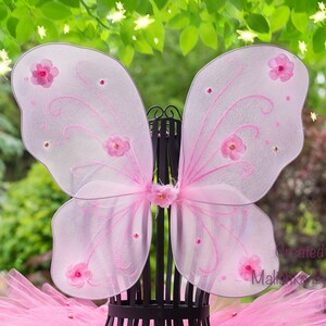 May include: Pink fairy wings with floral accents and a matching tutu. The wings are made of sheer fabric with pink glitter detailing and small pink flowers. The tutu is a fluffy, layered design. The text "Created by Malishka Boutique" is visible.