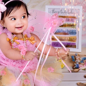 May include: A young girl dressed as a fairy with pink wings and a tutu, holding a wand with a pink ribbon. She is wearing a pink and gold dress with a pearl necklace and a gold bracelet. The background includes a sign with the text "things to do today" and a list of activities.