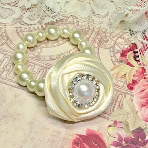 Puede incluir: Un brazalete de perlas blancas con un acento de flor de sat&eacute;n blanco. La flor tiene un centro de diamantes de imitaci&oacute;n con una perla en el medio.