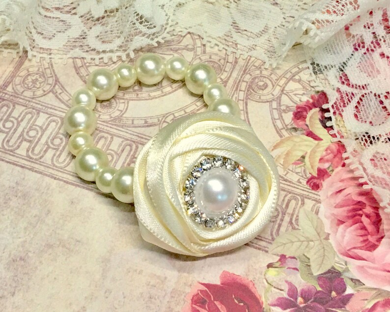Puede incluir: Un brazalete de perlas blancas con un acento de flor de sat&eacute;n blanco. La flor tiene un centro de diamantes de imitaci&oacute;n con una perla en el medio.