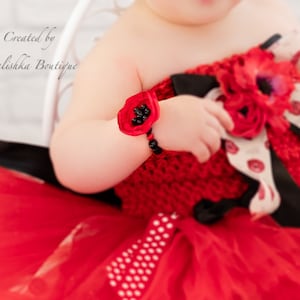 May include: A red and black baby dress with a poppy flower detail. The dress has a tulle skirt and a crocheted bodice. The baby is wearing a matching poppy flower bracelet.