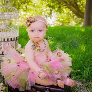 Puede incluir: Una ni&ntilde;a peque&ntilde;a con un tut&uacute; rosa y verde, una diadema de flores y un collar de perlas. Est&aacute; sentada en una maleta con una jaula de p&aacute;jaros en el fondo.