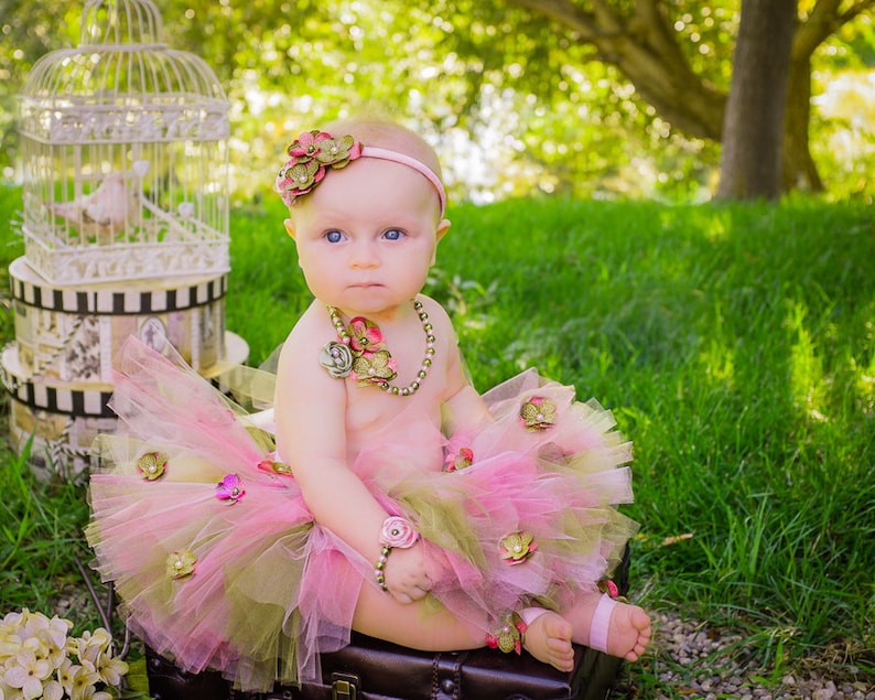 Puede incluir: Una ni&ntilde;a peque&ntilde;a con un tut&uacute; rosa y verde, una diadema de flores y un collar de perlas. Est&aacute; sentada en una maleta con una jaula de p&aacute;jaros en el fondo.