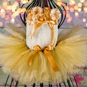 Op de afbeelding: Een gouden tule tutu met gouden glitteraccenten en een wit gehaakt topje met een gouden satijnen strik en een bloemversiering. De tekst "Gemaakt door Malishka Boutique" is zichtbaar in de rechterbenedenhoek van de afbeelding.