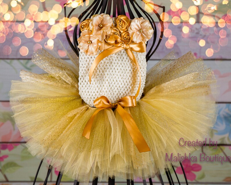 Op de afbeelding: Een gouden tule tutu met gouden glitteraccenten en een wit gehaakt topje met een gouden satijnen strik en een bloemversiering. De tekst "Gemaakt door Malishka Boutique" is zichtbaar in de rechterbenedenhoek van de afbeelding.