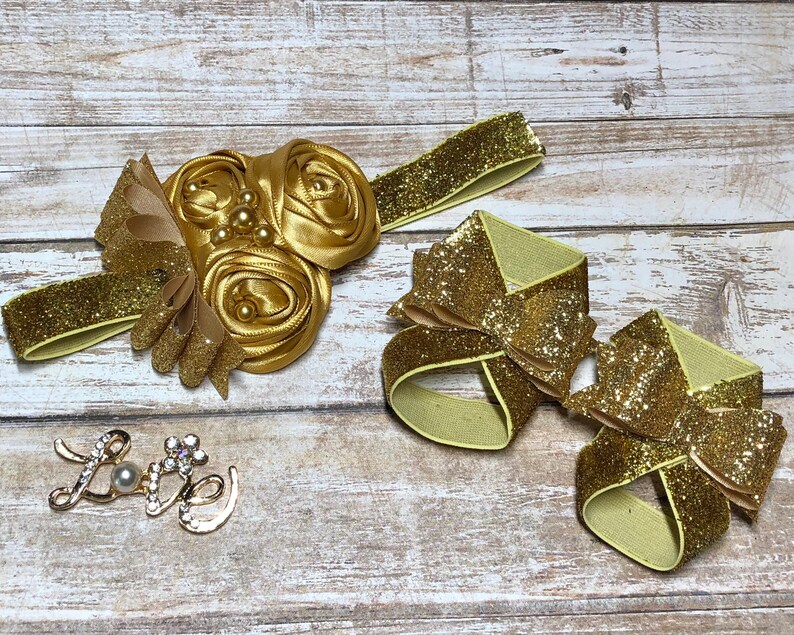 Op de afbeelding: Een set van twee gouden glitter haarbandjes met elastiekjes. De strikken zijn in de vorm van een bloem en een strik. Er is ook een gouden bedel met het woord "Love" en een parel in het midden.