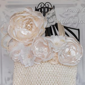 Puede incluir: Un vestido de beb&eacute; de crochet blanco con un gran adorno floral de sat&eacute;n blanco y cuentas de perlas. El vestido est&aacute; en un soporte negro con un borde de encaje blanco.