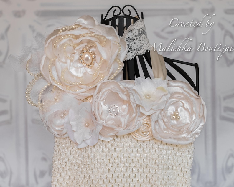 Puede incluir: Un vestido de beb&eacute; de crochet blanco con un gran adorno floral de sat&eacute;n blanco y cuentas de perlas. El vestido est&aacute; en un soporte negro con un borde de encaje blanco.