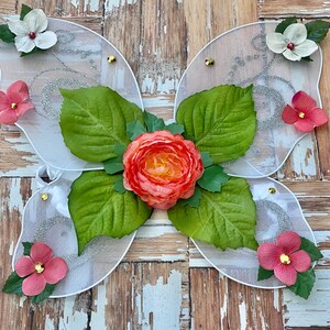 May include: White butterfly wings with a peach-colored flower and green leaves in the center. The wings are decorated with silver glitter and small white flowers.