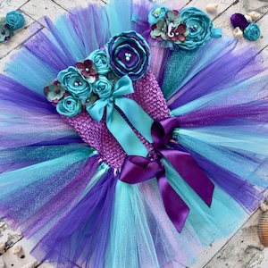 May include: A purple and blue tulle tutu with a purple crocheted bodice adorned with blue and purple fabric flowers. The bodice has a turquoise satin bow and a purple satin ribbon. The tutu is accented with glitter.