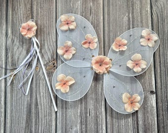 May include: A pair of white fairy wings with peach-colored flower accents. The wings are made of tulle and have a delicate, ethereal look. A matching wand with a peach-colored flower on the end is also included.