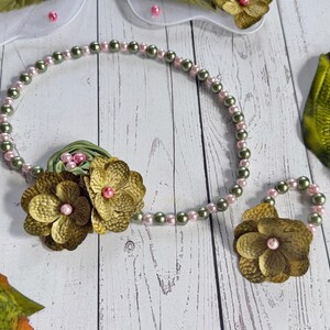 May include: A floral jewelry set featuring a necklace and a bracelet. The necklace and bracelet are made of alternating pink and olive green beads. The set includes two large, textured, gold-colored flowers with pink centers.