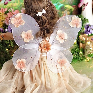 May include: A child wearing a white tulle dress with a pair of white butterfly wings decorated with pink flowers. The wings have a sparkly silver finish.