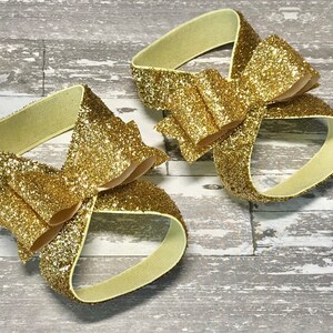 Op de afbeelding: Een paar gouden glitter enkelbandjes met een grote gouden glitter strik op elk bandje.
