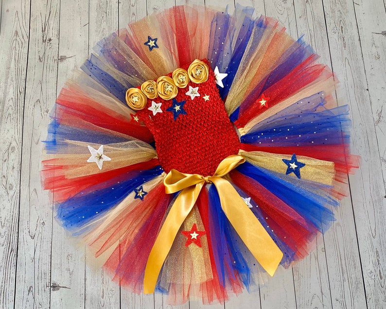 May include: Red, white, and blue tulle tutu with gold ribbon and gold stars. The tutu has a red crocheted top with gold flowers and white stars.