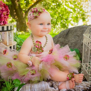 Puede incluir: Una ni&ntilde;a peque&ntilde;a con un tut&uacute; rosa y verde con adornos de flores, una diadema de flores y un collar de perlas. Est&aacute; sentada en una manta con una jaula de p&aacute;jaros y una caja en el fondo.