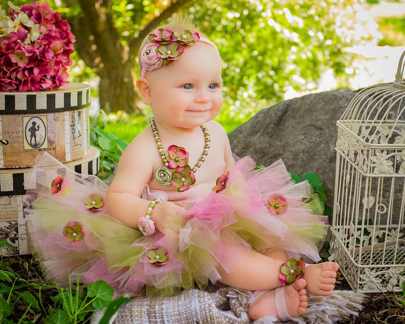 Puede incluir: Una ni&ntilde;a peque&ntilde;a con un tut&uacute; rosa y verde con adornos de flores, una diadema de flores y un collar de perlas. Est&aacute; sentada en una manta con una jaula de p&aacute;jaros y una caja en el fondo.