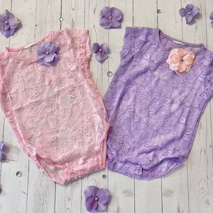 May include: Two lace baby bodysuits, one pink and one purple, with delicate floral designs. Both bodysuits have a round neckline and short sleeves.