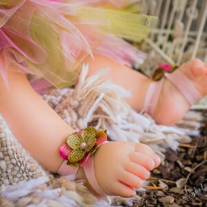 Puede incluir: Los pies de un beb&eacute; con brazaletes de tobillo de flores rosas y verdes con lazos de cinta rosa. El beb&eacute; lleva una falda de tul rosa y verde.