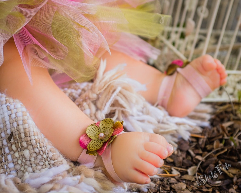 Puede incluir: Los pies de un beb&eacute; con brazaletes de tobillo de flores rosas y verdes con lazos de cinta rosa. El beb&eacute; lleva una falda de tul rosa y verde.