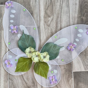 May include: White fairy wings with green leaves and purple flowers. The wings are made of sheer fabric and have a sparkly finish.