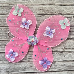May include: Pink butterfly wings with a sheer fabric design, adorned with fabric flowers in shades of purple, light blue, and pink. Silver glitter accents and small, colorful gems add sparkle. The wings have a delicate, whimsical appearance.