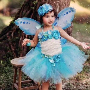 May include: A young girl wearing a blue and white fairy costume with a tulle skirt, butterfly wings, and a flower crown. She is holding a flower in her hand and is standing in front of a wooden stool.