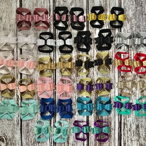 Op de afbeelding: Een verzameling van 24 paar glinsterende babysandalen in verschillende kleuren en strikdesigns. De sandalen zijn gemaakt van elastiek en hebben een strik aan de voorkant.