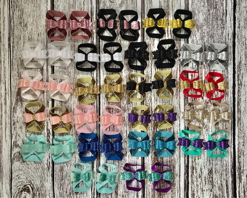 Op de afbeelding: Een verzameling van 24 paar glinsterende babysandalen in verschillende kleuren en strikdesigns. De sandalen zijn gemaakt van elastiek en hebben een strik aan de voorkant.