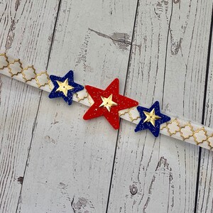 May include: A white headband with a gold geometric pattern and three star-shaped embellishments. The stars are red, blue, and blue with gold glitter.