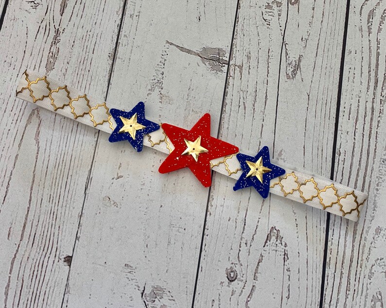 May include: A white headband with a gold geometric pattern and three star-shaped embellishments. The stars are red, blue, and blue with gold glitter.