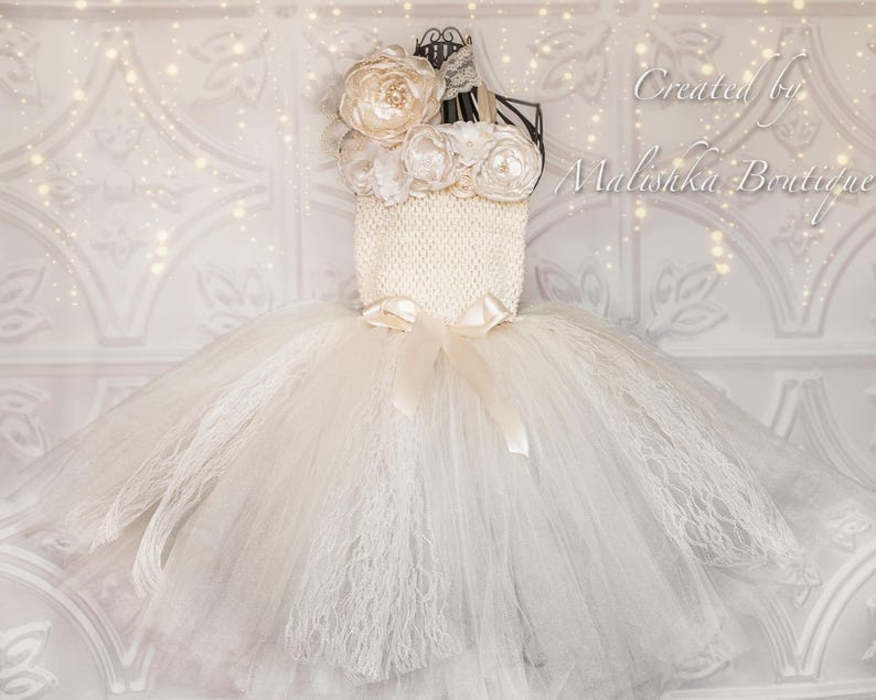 Puede incluir: Un tut&uacute; de tul blanco con un corpi&ntilde;o de ganchillo blanco y un adorno de flores blancas. El corpi&ntilde;o tiene una cinta de sat&eacute;n atada alrededor de la cintura. Creado por Malishka Boutique.