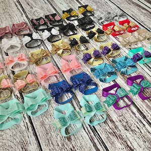 Op de afbeelding: Een verscheidenheid aan glinsterende baby-barefoot sandalen met strikken in verschillende kleuren, waaronder roze, blauw, groen, paars en goud.