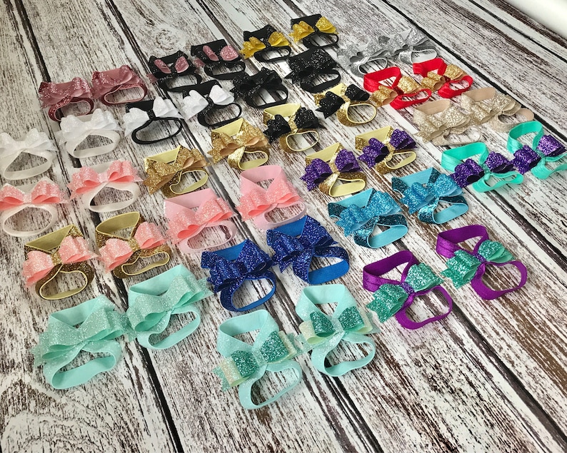 Op de afbeelding: Een verscheidenheid aan glinsterende baby-barefoot sandalen met strikken in verschillende kleuren, waaronder roze, blauw, groen, paars en goud.