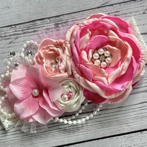 May include: A pink and white flower headband with pearl accents. The headband is made of lace and tulle and features a large pink flower with a smaller pink flower and a white flower.