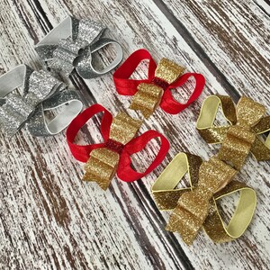 Op de afbeelding: Vijf glitterbogen in verschillende kleuren, waaronder zilver, goud en rood. De strikken zijn vastgemaakt met elastiekjes en zijn perfect voor haaraccessoires.