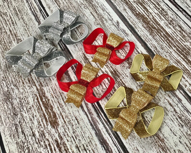 Op de afbeelding: Vijf glitterbogen in verschillende kleuren, waaronder zilver, goud en rood. De strikken zijn vastgemaakt met elastiekjes en zijn perfect voor haaraccessoires.