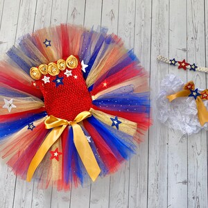 May include: A red, white, and blue tulle tutu with gold stars and a red bodice with gold and white stars. A white lace diaper cover with a gold bow and a gold headband with red and blue stars.