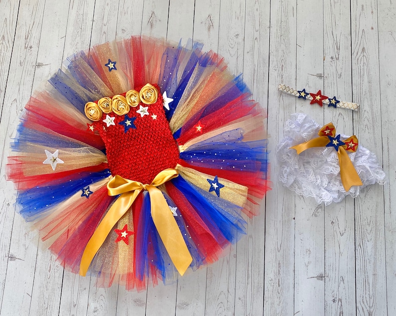 May include: A red, white, and blue tulle tutu with gold stars and a red bodice with gold and white stars. A white lace diaper cover with a gold bow and a gold headband with red and blue stars.