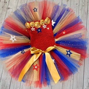May include: A red, white, and blue tulle tutu with gold glitter and stars. The tutu has a red crocheted top with gold and white flower embellishments.