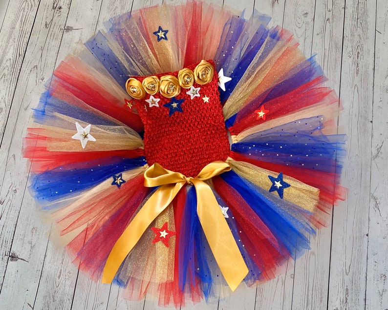 May include: A red, white, and blue tulle tutu with gold glitter and stars. The tutu has a red crocheted top with gold and white flower embellishments.