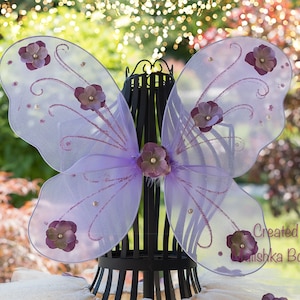 May include: Purple fairy wings with a sparkly lavender tulle fabric and purple flower embellishments. The wings are displayed on a black stand. Created by Mishka Boutique.