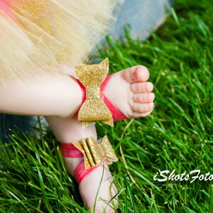 Op de afbeelding: Een paar babysandalen met rode bandjes en gouden glitter strikjes. De sandalen liggen op een bed van groen gras.