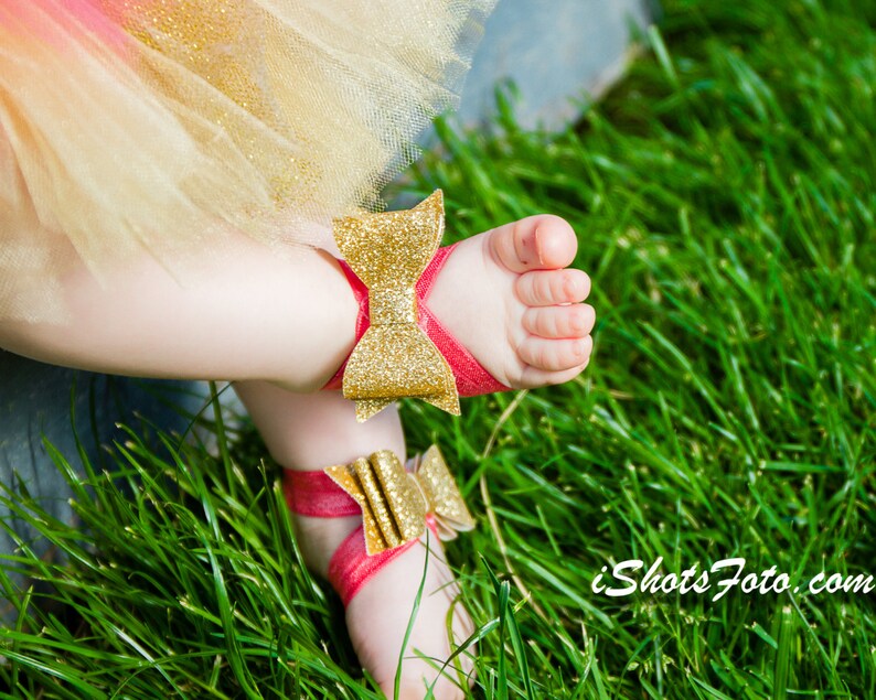 Op de afbeelding: Een paar babysandalen met rode bandjes en gouden glitter strikjes. De sandalen liggen op een bed van groen gras.